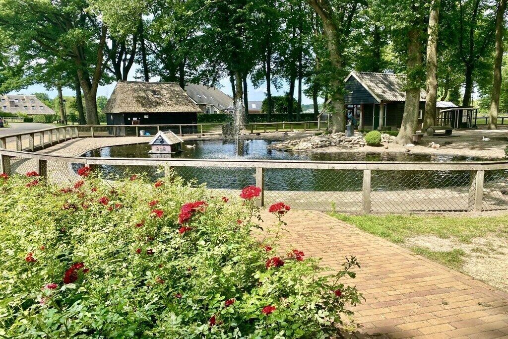 A pond with a wooden bridge, surrounded by trees and a thatched-roof building.