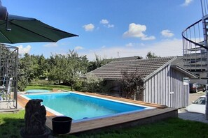A swimming pool with a wooden deck, a stone statue, and a covered patio area.