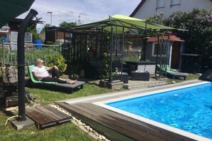 A backyard with a swimming pool, a covered patio area, and a person relaxing on a lounge chair. - In the Old Schoolhouse (Schrozberg)