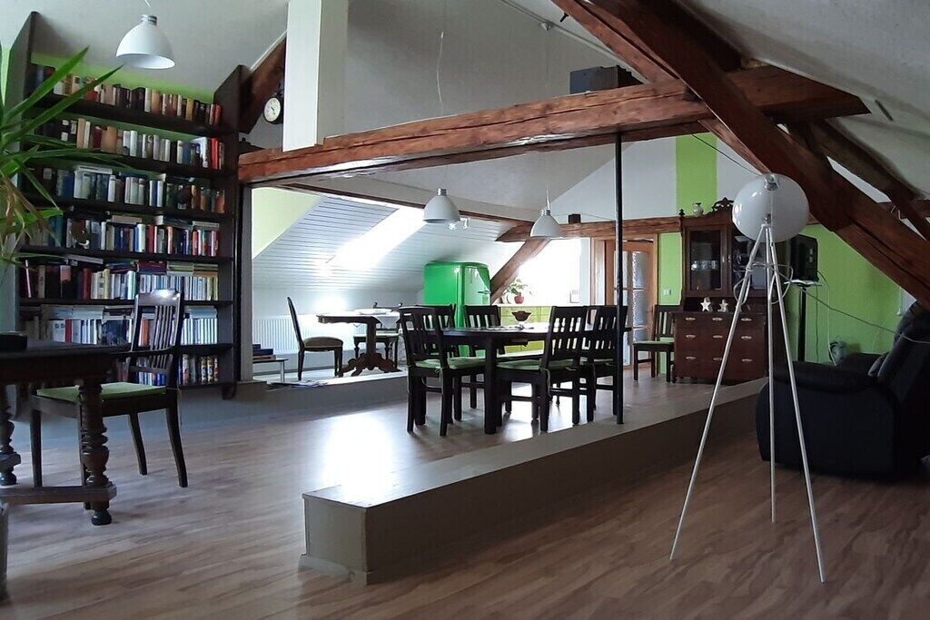 Ein geräumiges Loft mit hoher Decke, freiliegenden Holzbalken, einem Essbereich mit langem Tisch und Stühlen, einem Bücherregal voller Bücher und einem grünen Sessel.