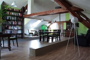 Ein geräumiges Loft mit hoher Decke, freiliegenden Holzbalken, einem Essbereich mit langem Tisch und Stühlen, einem Bücherregal voller Bücher und einem grünen Sessel.