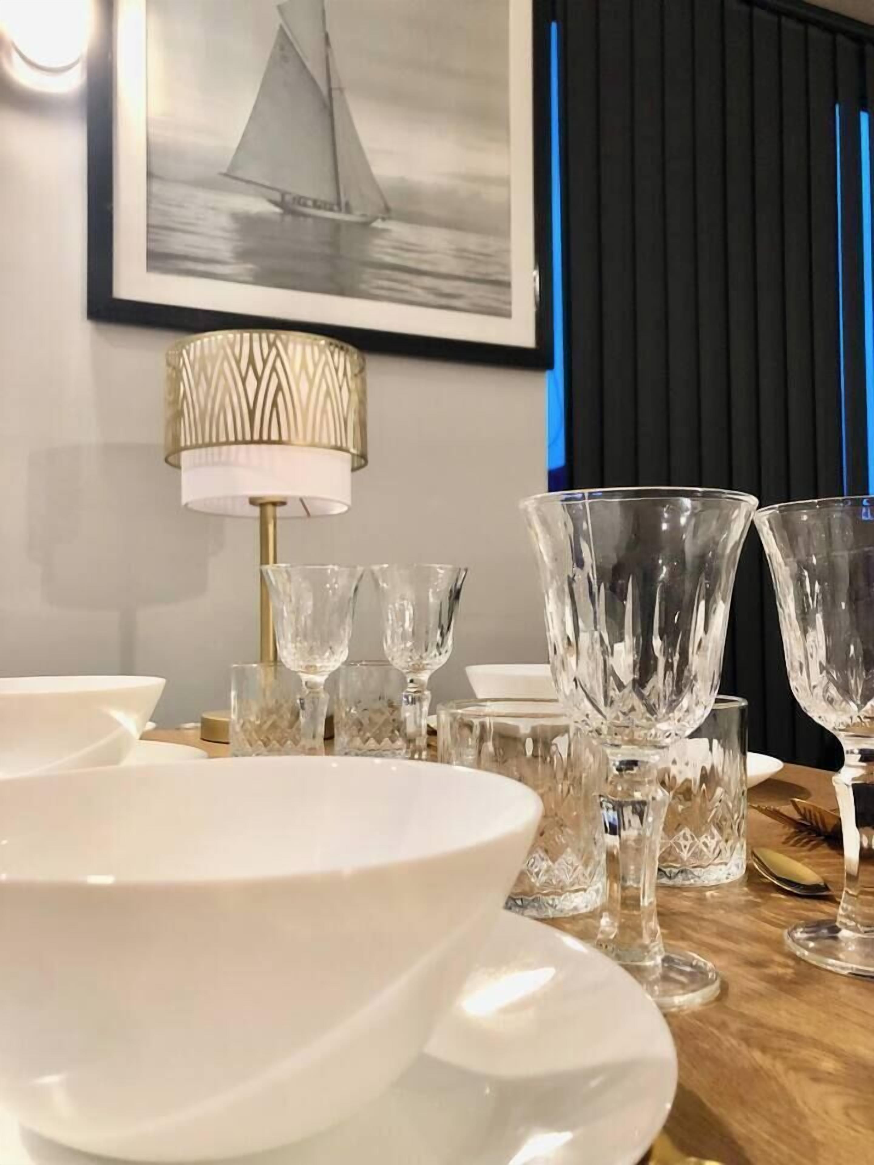 Taula de menjador amb plats blancs i vidrieria de cristall, una làmpada decorativa i un quadre amb un veler a la paret.