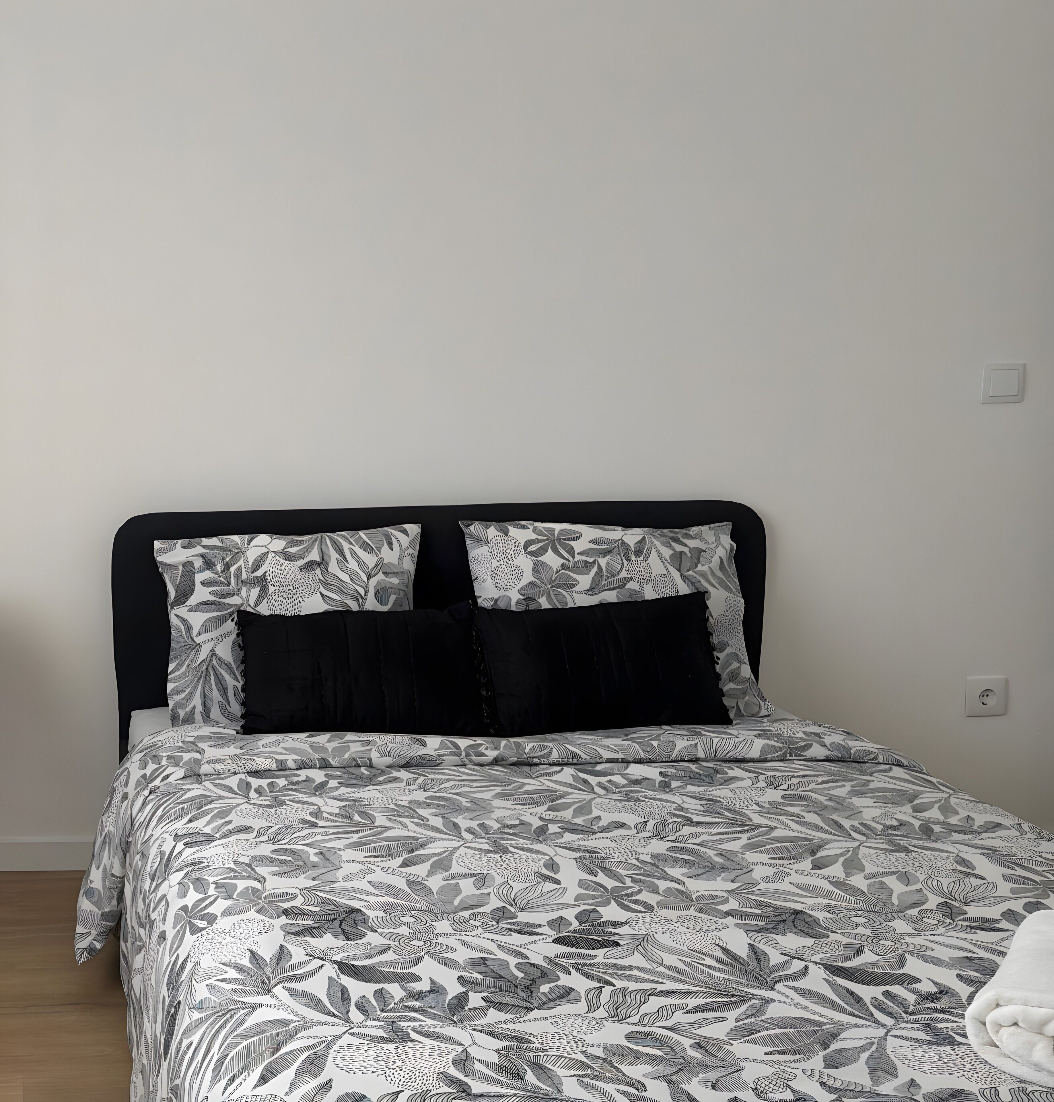 Un lit avec une couvre-lit à motifs et deux oreillers, une tête de lit noire et un mur uni avec une prise de courant.