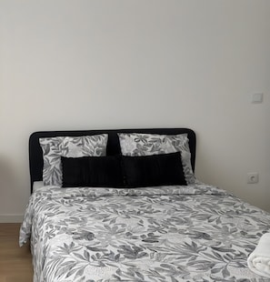 A bed with a patterned bedspread and two pillows, a black headboard, and a plain wall with a power outlet.