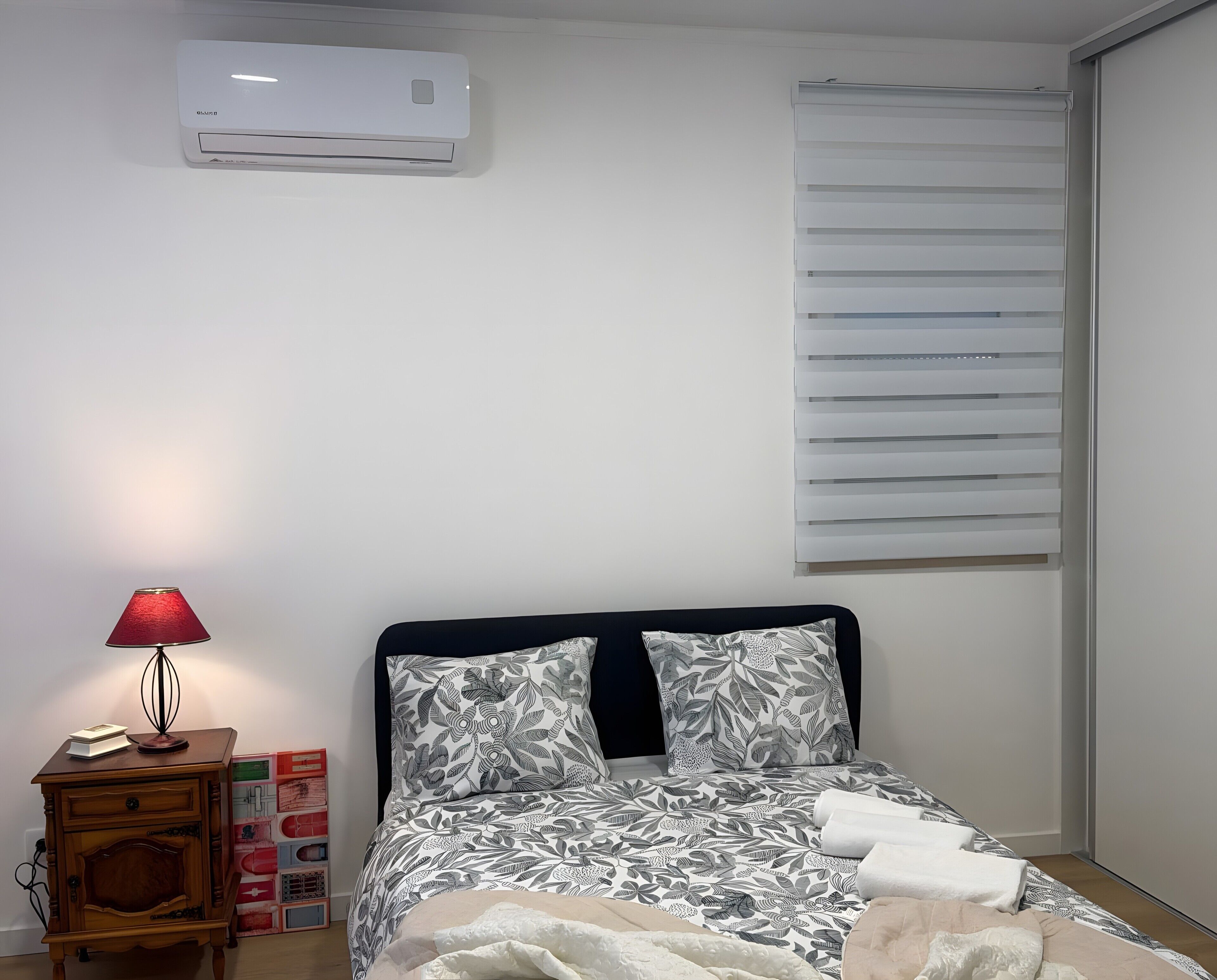 A bedroom with a bed, a bedside table with a lamp, a window with blinds, and an air conditioning unit on the wall.