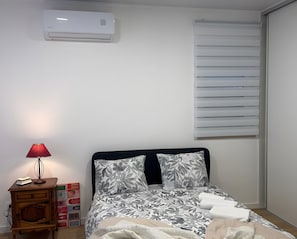 A bedroom with a bed, a bedside table with a lamp, a window with blinds, and an air conditioning unit on the wall. - Apartment 'Mira D'Aire Serenity House Confort' (Mira de Aire)