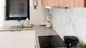 Une cuisine moderne avec des meubles blancs, une plaque de cuisson à induction noire, un four encastré et une fenêtre avec des stores.