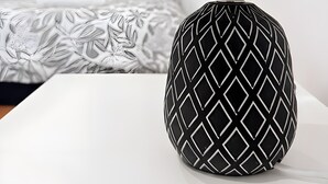 Une lampe ornée d’un motif géométrique sur un support blanc, un lit avec une literie à motifs et un mur uni en arrière-plan.