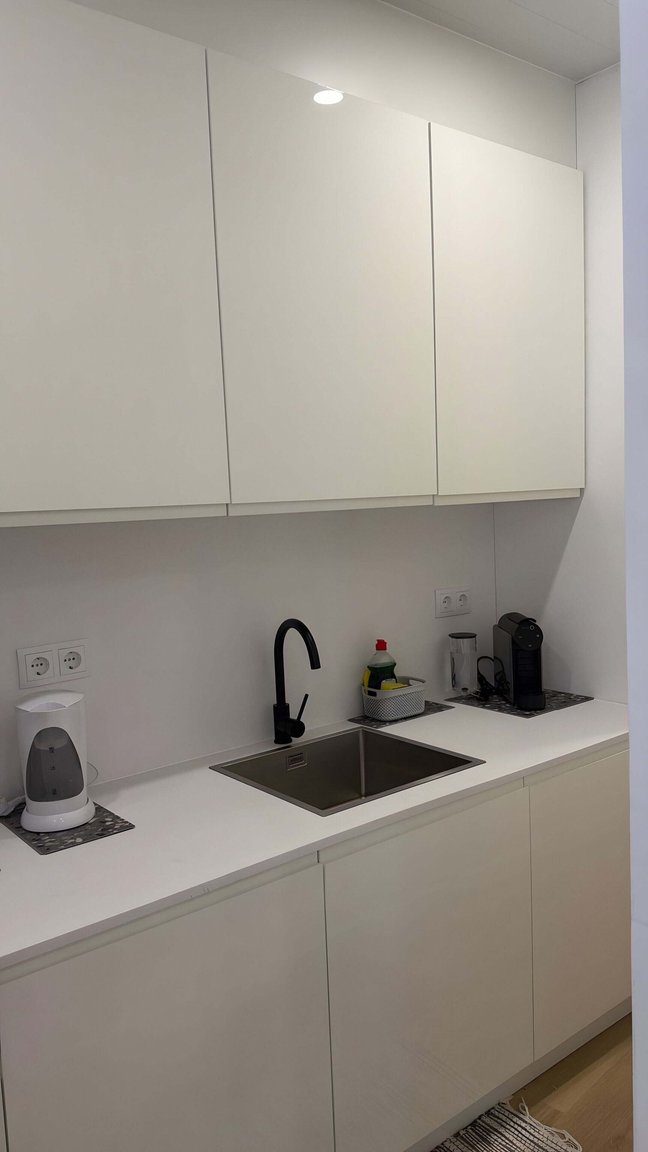 Une cuisine moderne avec des meubles blancs, un évier en acier inoxydable, une bouilloire, une machine à café et un petit panier contenant des produits de nettoyage.
