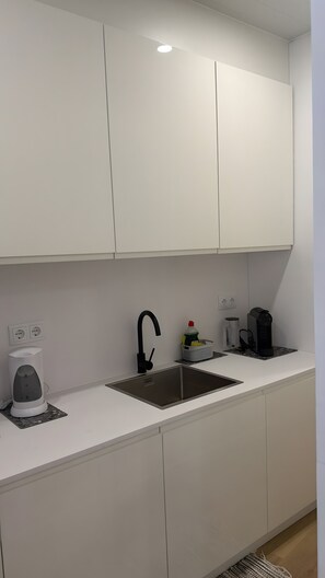 A modern kitchen with white cabinetry, a stainless steel sink, a kettle, a coffee maker, and a small basket with cleaning supplies.