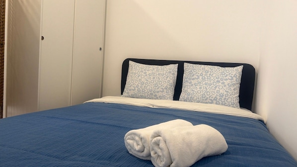 Un lit simple avec une couvre-lit bleue et deux oreillers à motifs floraux. Une serviette blanche est soigneusement pliée et posée sur le lit.