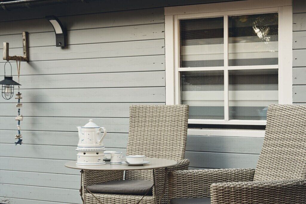 A small outdoor seating area with wicker chairs and a table, a white teapot, and a window with blinds.