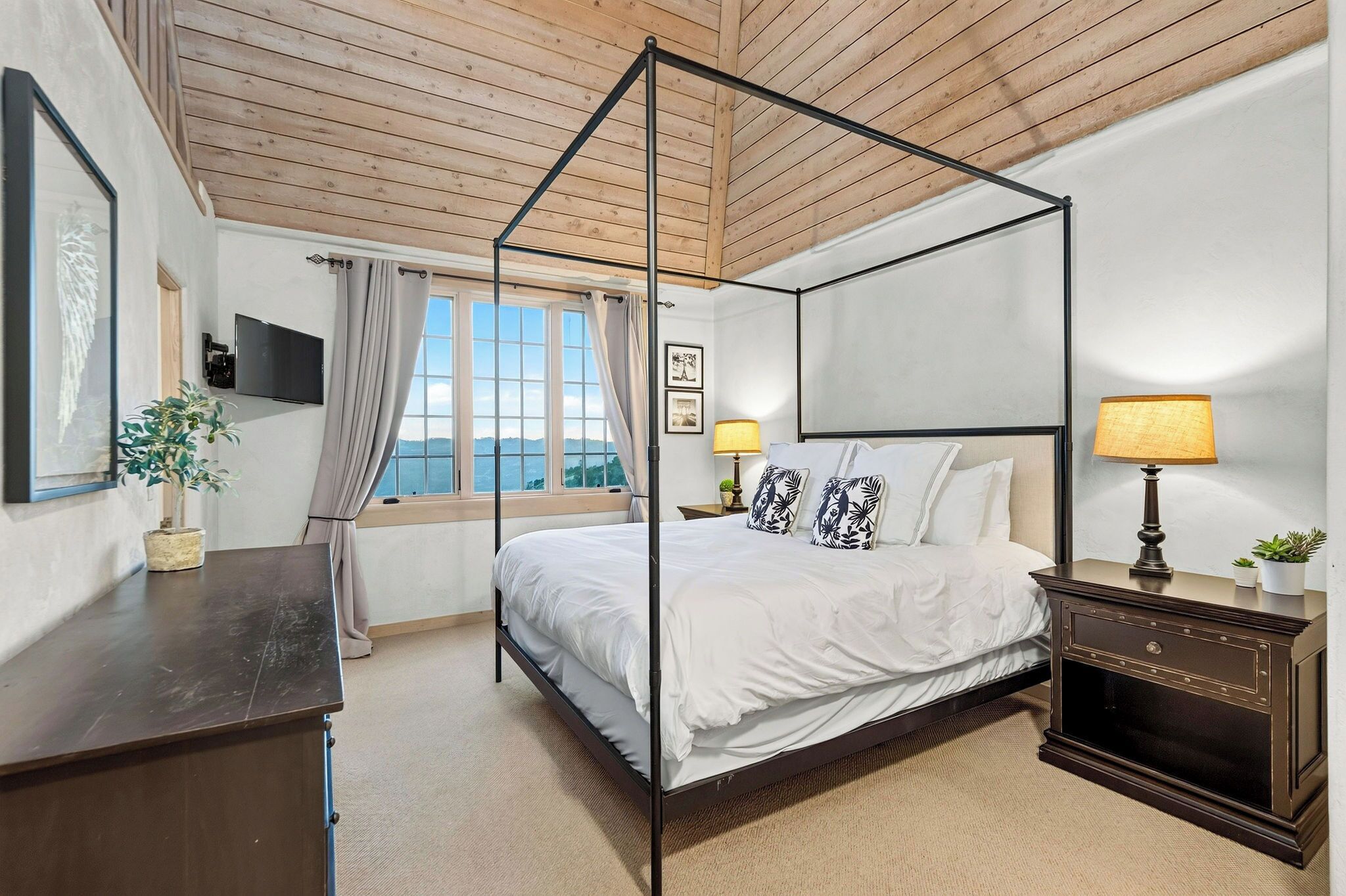 Una cama con dosel con ropa blanca, cabecera de madera y vista al mar a través de un ventanal grande.
