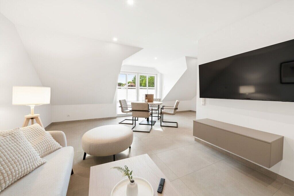 Ein modernes Wohnzimmer mit einer Couch, Hocker und Couchtisch. Es gibt einen Essbereich mit Stühlen und einem Tisch, und ein an der Wand befestigtes Fernsehgerät.