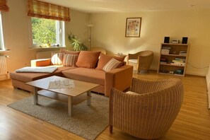 A living room with a sofa, a coffee table, a wicker chair, a bookshelf, and a window with curtains. - Holiday apartment with 1 bedroom (Osterode - Lerbach)