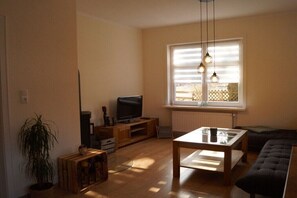 Ein Wohnzimmer mit Fernseher, Holzmöbeln, einer Topfpflanze und einem Fenster mit Jalousien.