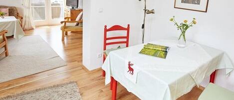 Una habitación con una mesa de comedor preparada para dos personas, una silla roja, un mantel blanco, un libro, un jarrón con flores y un televisor al fondo.