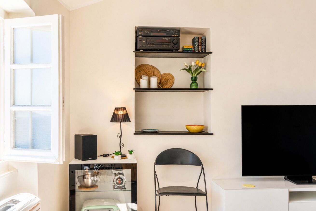 A modern living room with a flat-screen TV, a black chair, a white cabinet, and a shelf with books and decorative items.