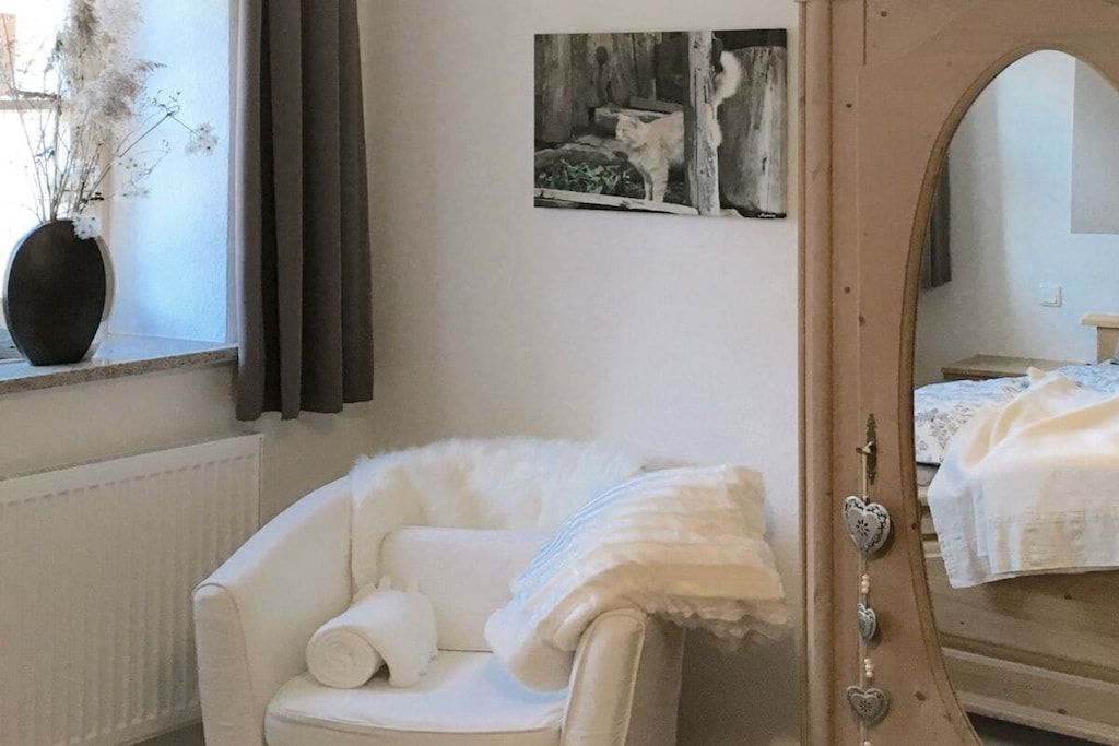 A white armchair with a rolled-up pillow, a framed picture on the wall, and a mirror with a heart-shaped decoration.