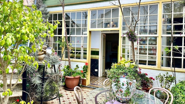 En innergård med ett glasbord, krukväxter och en byggnad med stora fönster och en grön fasad.