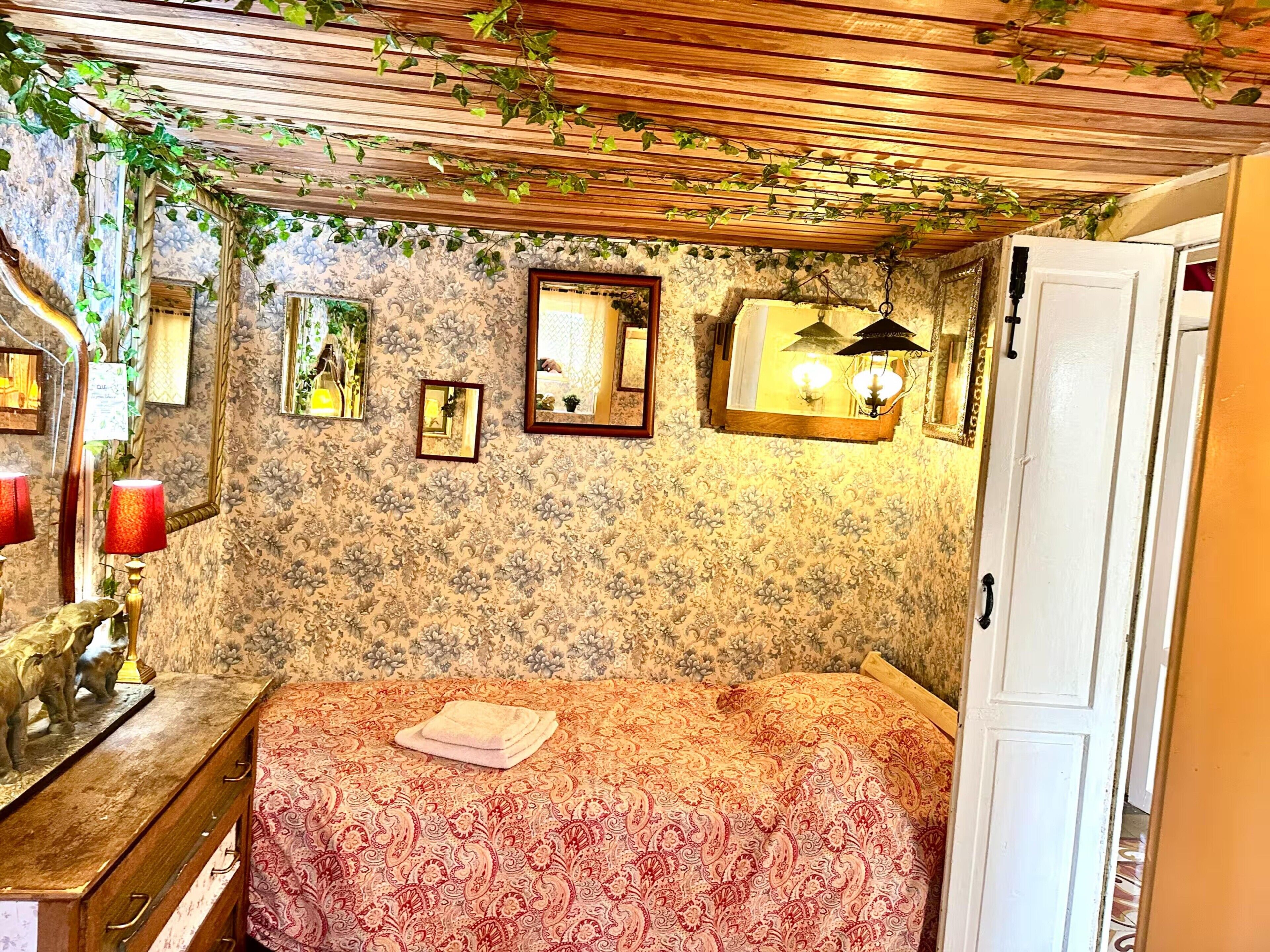 Une pièce avec un papier peint à motifs floraux, un lit recouvert d’une couvre-lit à motifs, une commode en bois et un miroir.