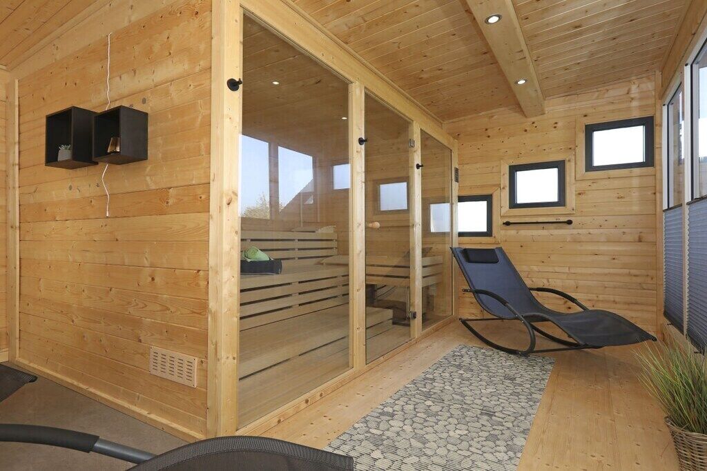 A wooden cabin interior with a sliding glass door leading to a balcony, a black lounge chair, and a small shelf with items.