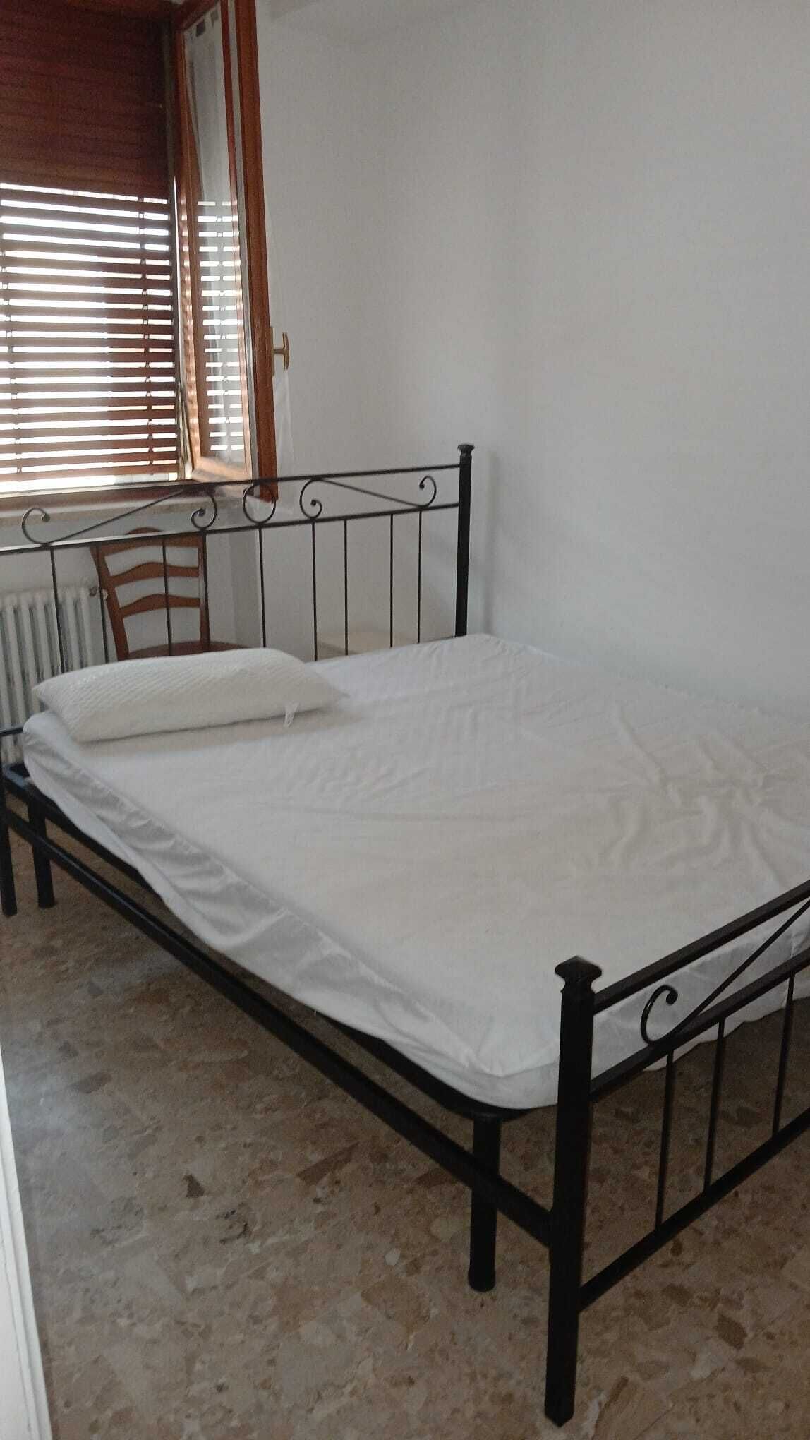 Ein Einzelbett mit Metallgestell in einem Zimmer mit Heizkörper und einem Fenster mit Jalousien.