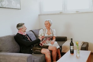 Two people seated on a grey sofa in a room with a window, a table with a bottle and a glass, and a vase with a flower. - Holiday apartment in Großklein with sauna (Großklein)