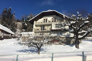 Ein zweistöckiges Gebäude mit einem Balkon, umgeben von schneebedeckten Bäumen und einer Steinmauer.