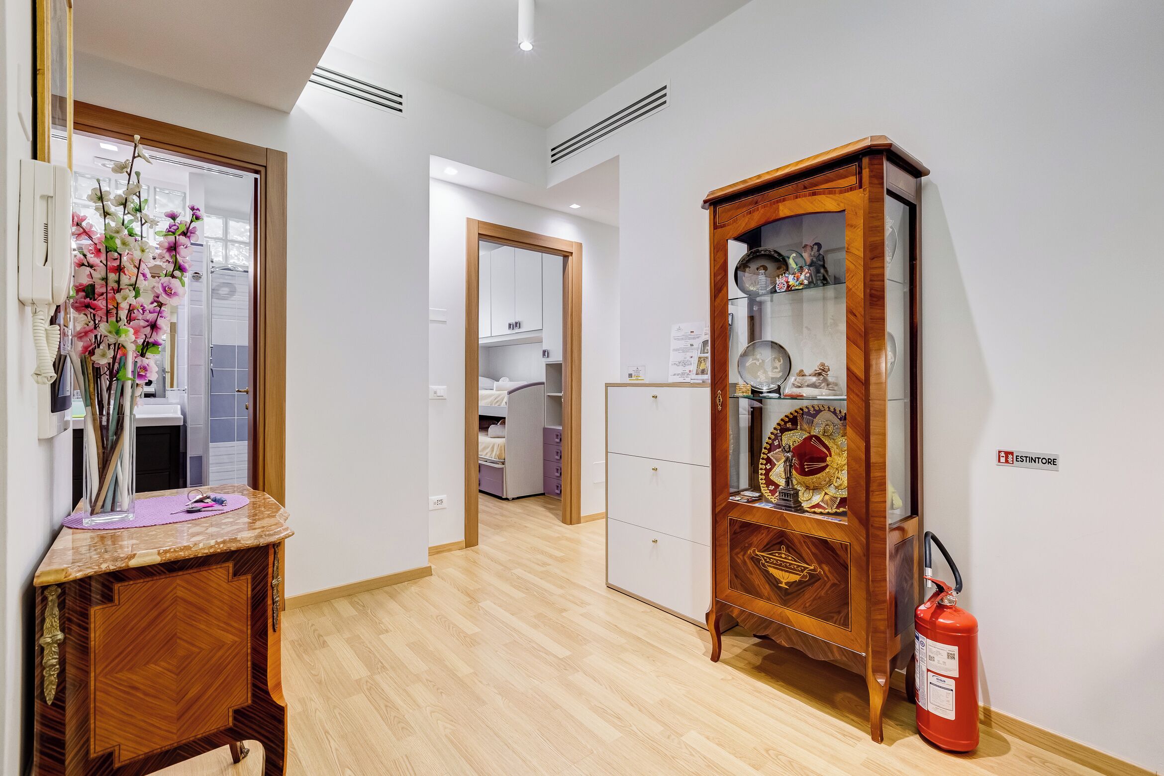A hallway with a wooden cabinet displaying decorative items, a fire extinguisher, and a mirror with a floral arrangement.