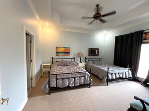 Ein Schlafzimmer mit zwei Betten, einem Deckenventilator, einem Gemälde, einem Nachttisch mit Lampe und einem Fenster mit Vorhängen.