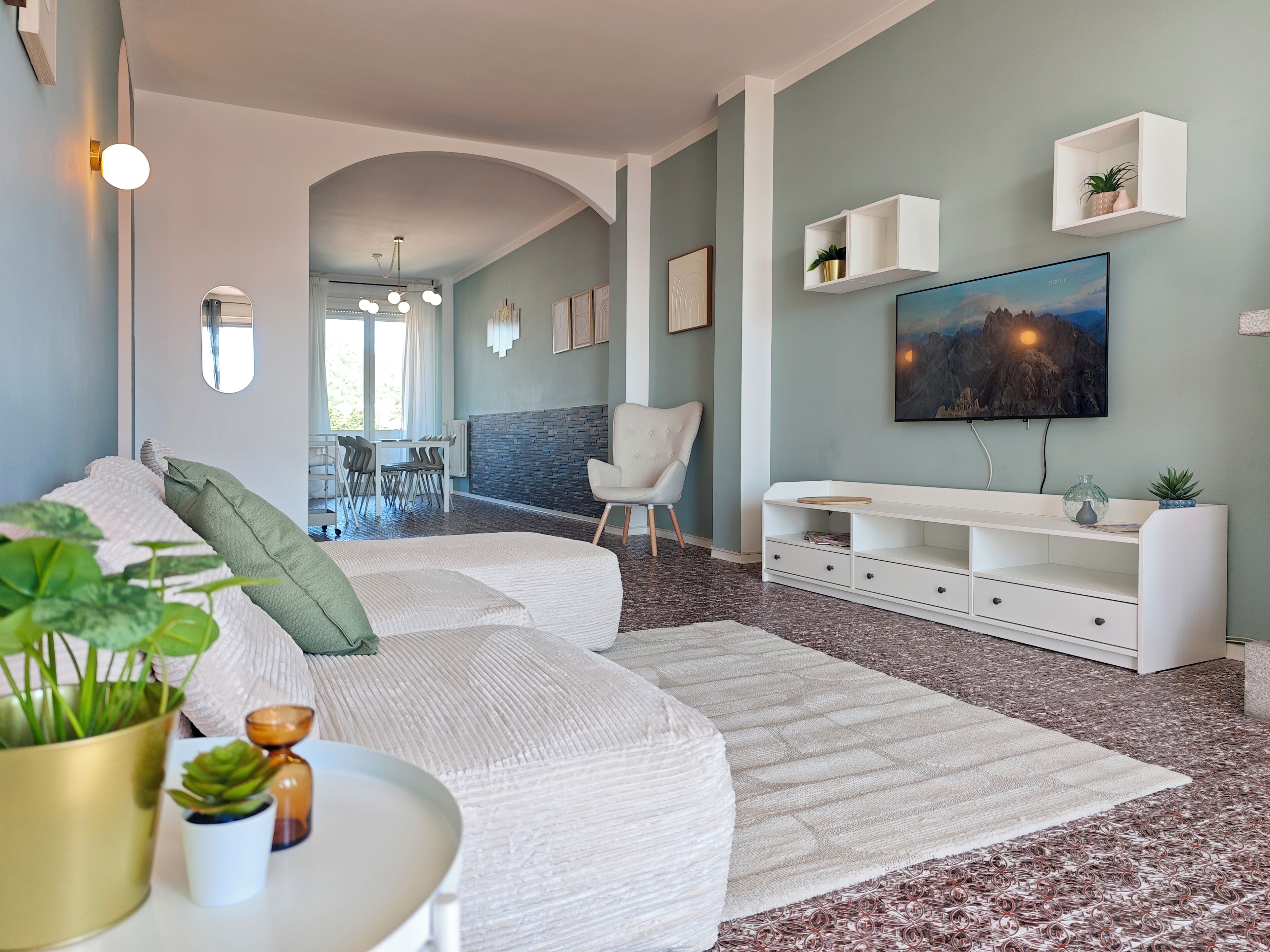 Een moderne woonkamer met een hoekbank, een eethoek en een wandtelevisie.