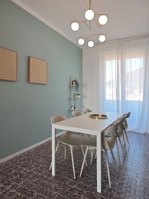 Een eetgedeelte met een witte tafel en stoelen, een plafondlamp en een raam met gordijnen.