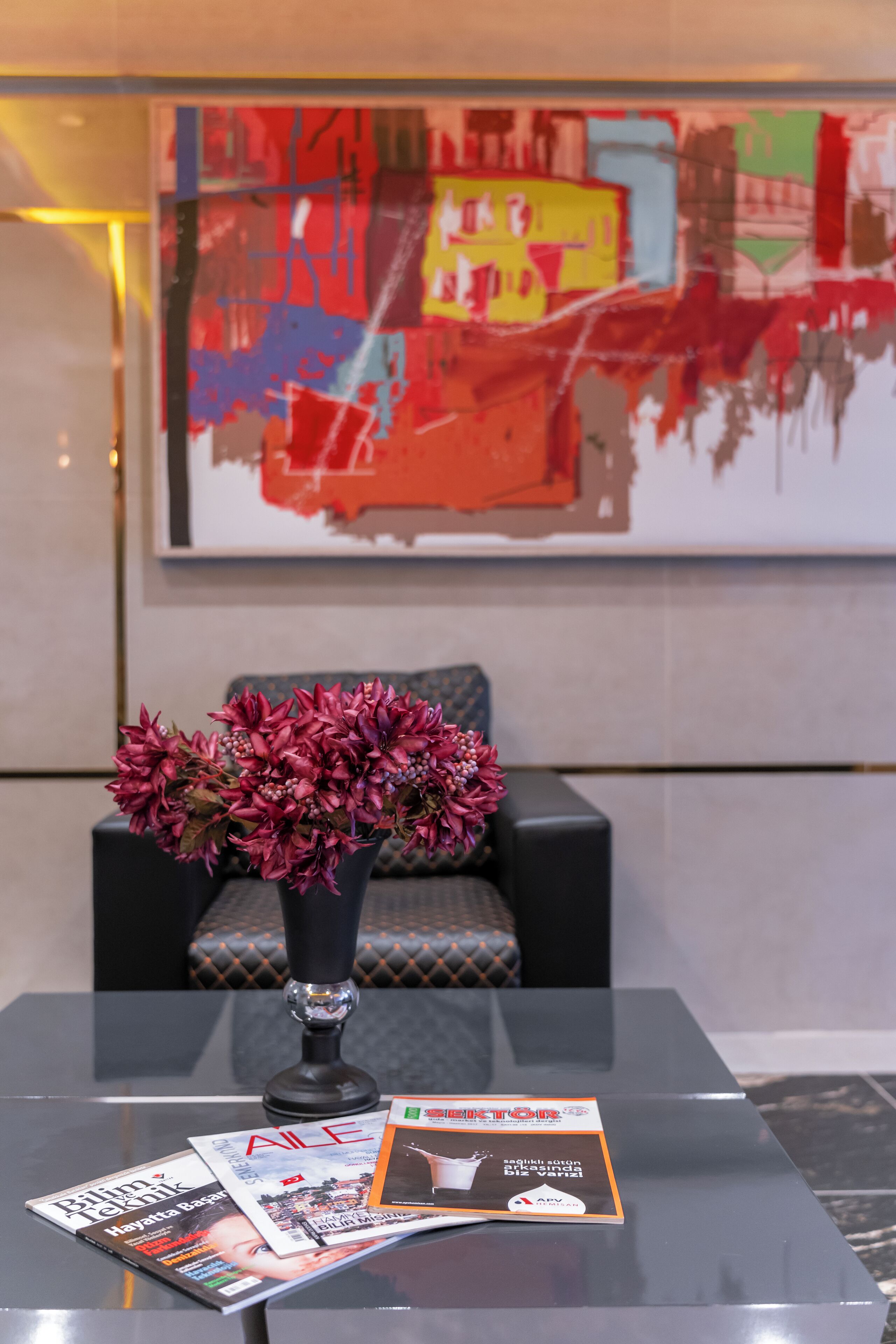 Um lobby moderno com uma mesa de centro de vidro, um vaso de flores e revistas. Uma pintura abstrata colorida está na parede atrás de um sofá.