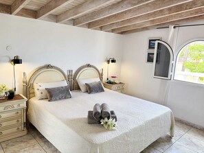 Ein Doppelbett mit weißer, strukturierter Bettdecke, zwei Nachttische mit Lampen, ein Fenster mit Blick auf Grünflächen und eine Holzbalkendecke.