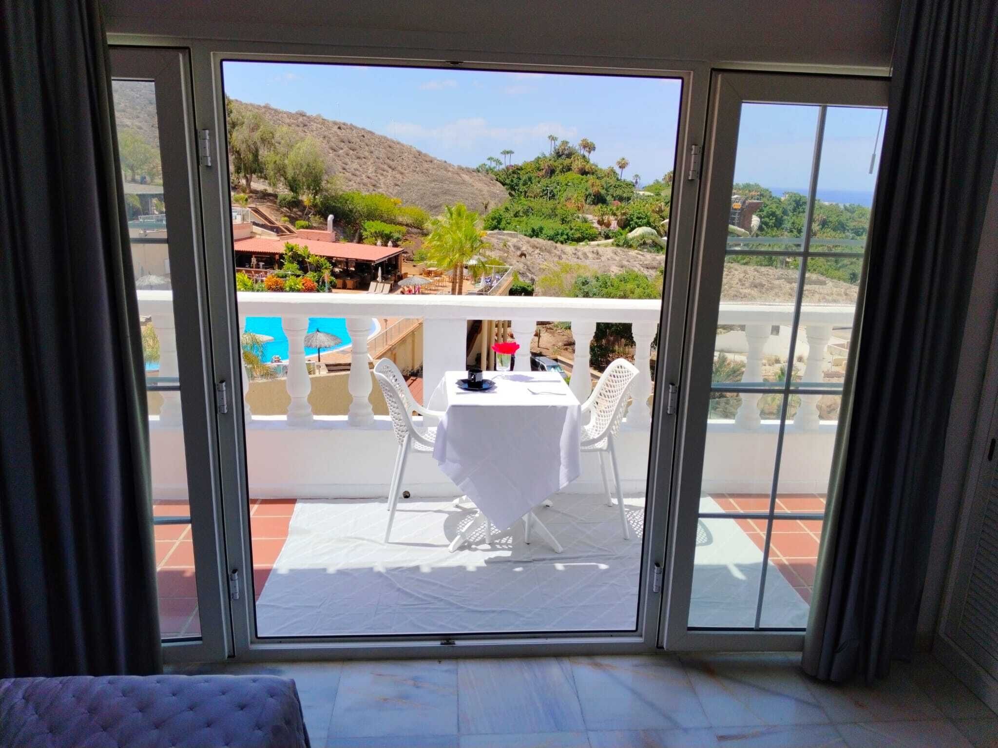 A room with a sliding glass door leading to an outdoor terrace with a table and chairs, a pool, and a view of a hillside.