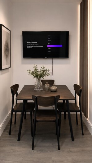 A dining area with a table, chairs, a vase with plants, and a wall-mounted television displaying a language selection menu. - 3 Bedroom House Sleeps 6 -garden -private Parking (Feltham)