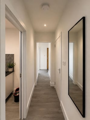 A hallway with a wooden floor, white walls, and a large mirror on the right. - 3 Bedroom House Sleeps 6 -garden -private Parking (Feltham)