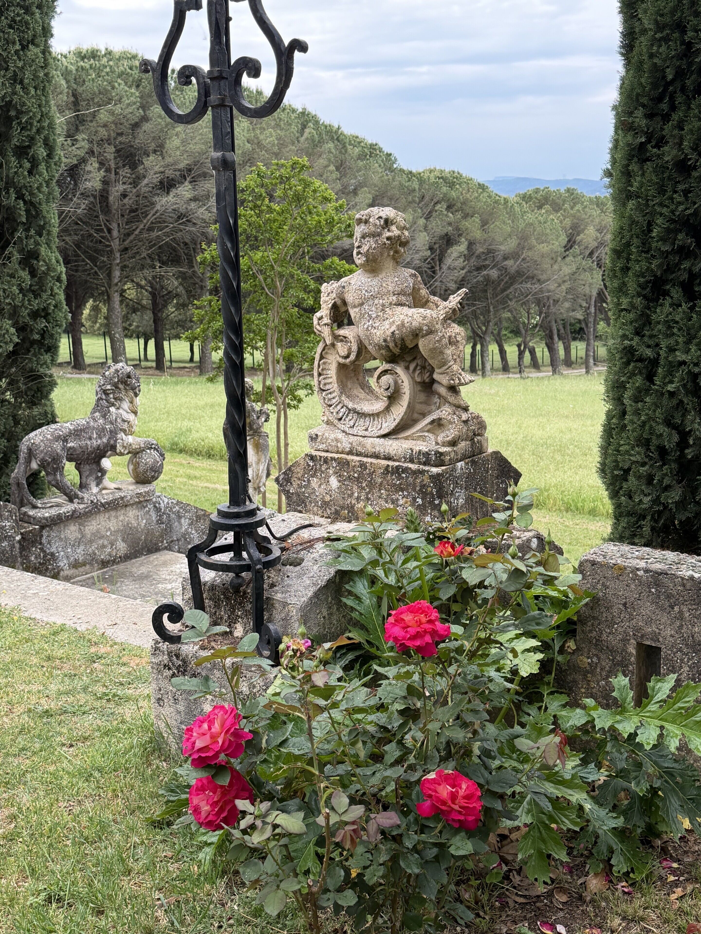 Un jardín con una estatua de piedra de una figura sosteniendo una planta, rodeado de rosales y un farol de metal.