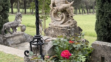 Un jardín con una estatua de piedra de una figura sosteniendo una planta, rodeado de rosales y un farol de metal.
