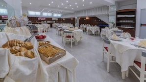 Une grande salle à manger avec de nombreuses tables dressées pour les repas, un espace buffet proposant des pâtisseries et du pain, et un escalier menant à un niveau supérieur.
