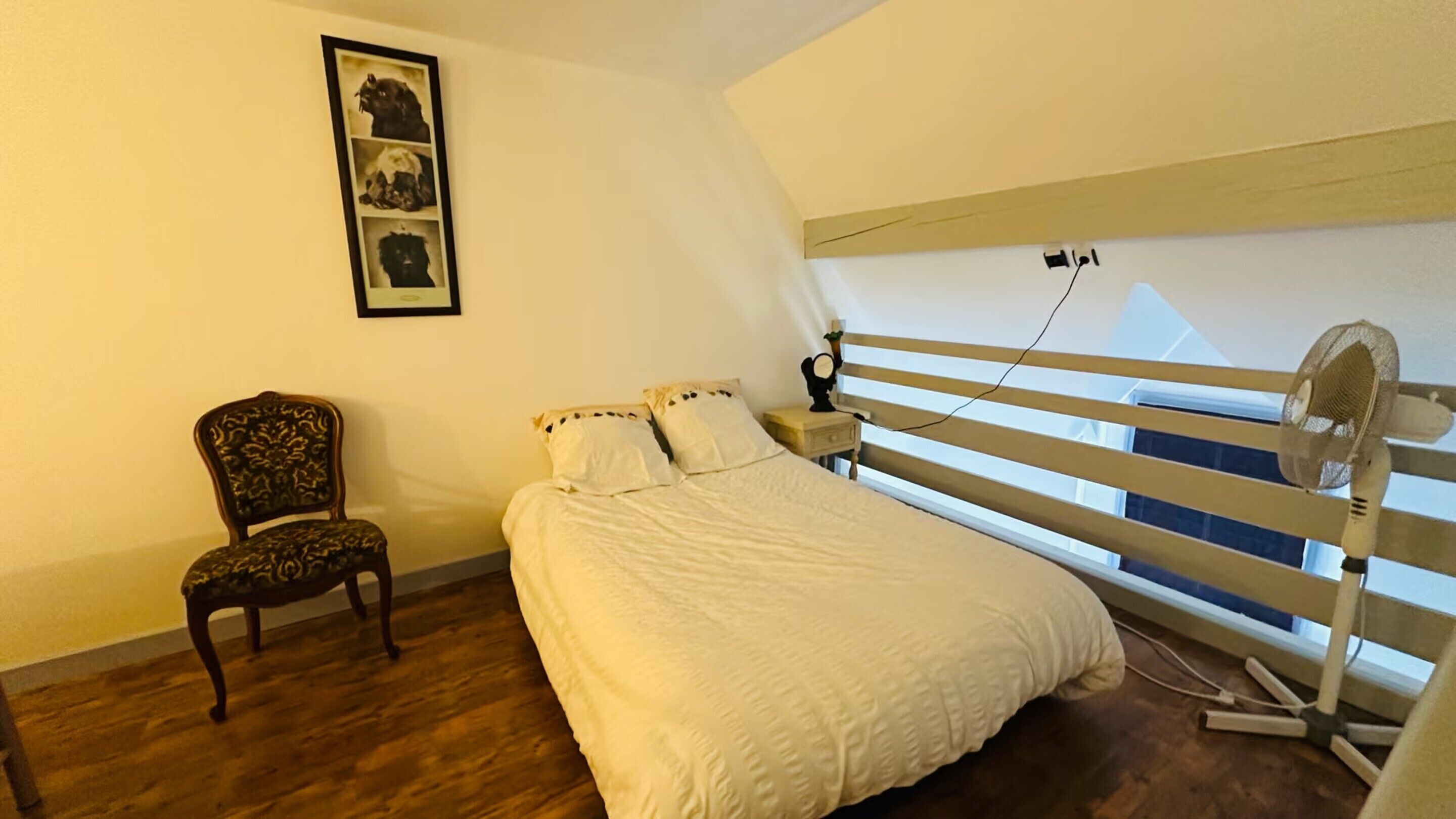 A bedroom with a bed, a chair, a window with a view, and a wall-mounted fan.