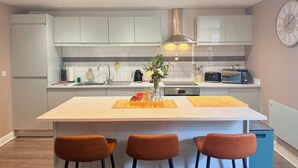 Una cocina moderna con armarios blancos, una isla central con taburetes, un reloj en la pared e iluminación colgante.