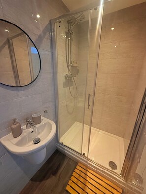 Ein modernes Badezimmer mit einer gläsernen Duschkabine, einem Waschbecken mit Spiegel und einer Holzmatte im Eingangsbereich.
