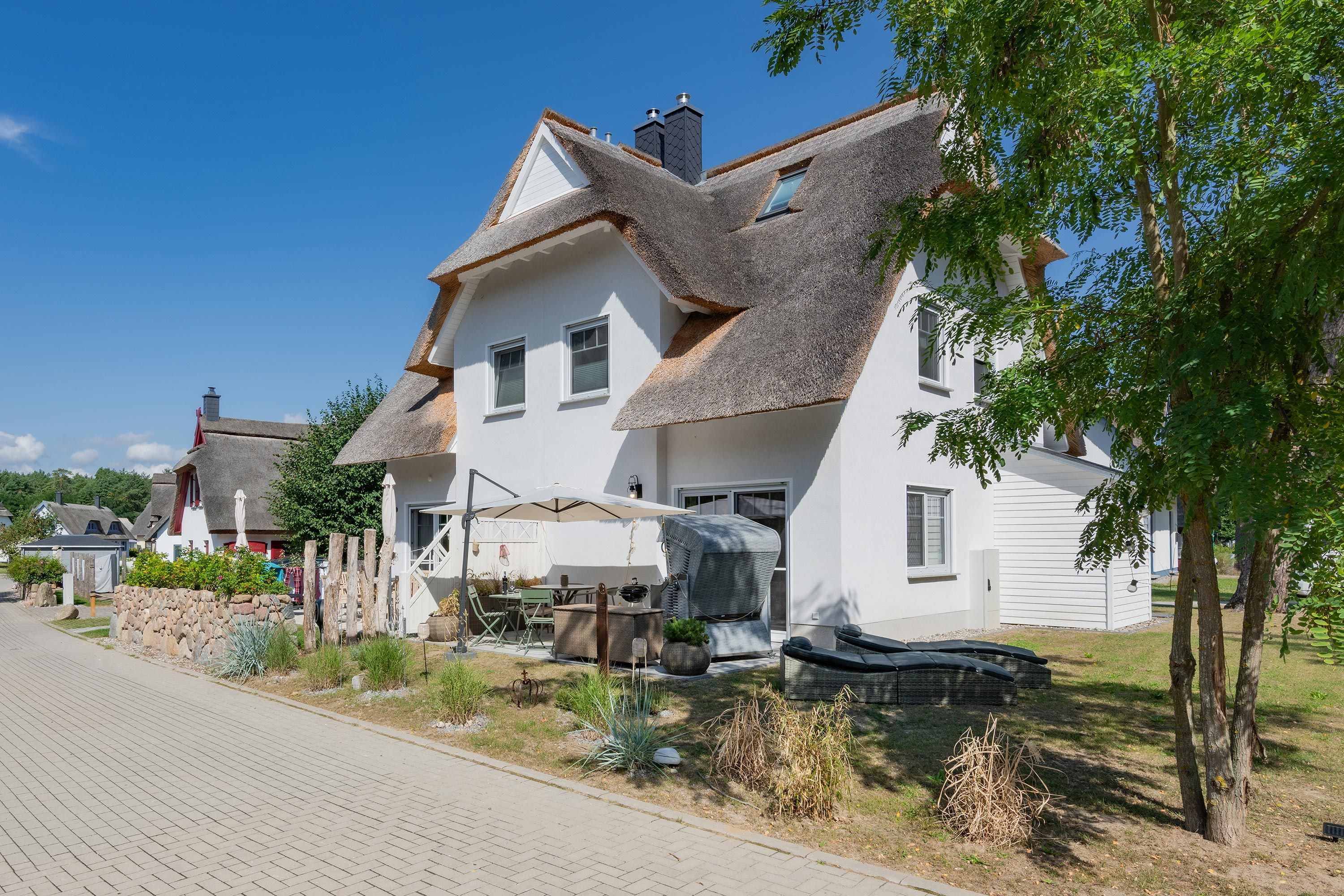 A white house with a thatched roof, a paved driveway, and a garden with various plants.