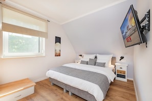 A bedroom with a bed, bedside tables, a nightstand, a window with blinds, and a wall-mounted TV. - LYKKE LUNA Vacation Home with Sauna and Fireplace on Usedom (Zirchow)