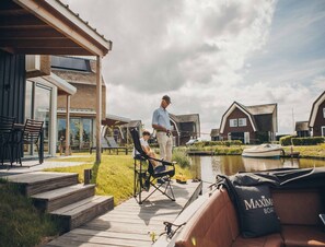 Ein modernes Haus mit einer Terrasse mit Blick auf einen Kanal, ein Boot liegt am Kai, und eine Person beim Angeln.