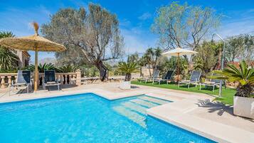 A swimming pool with lounge chairs and umbrellas, surrounded by trees and a clear sky.