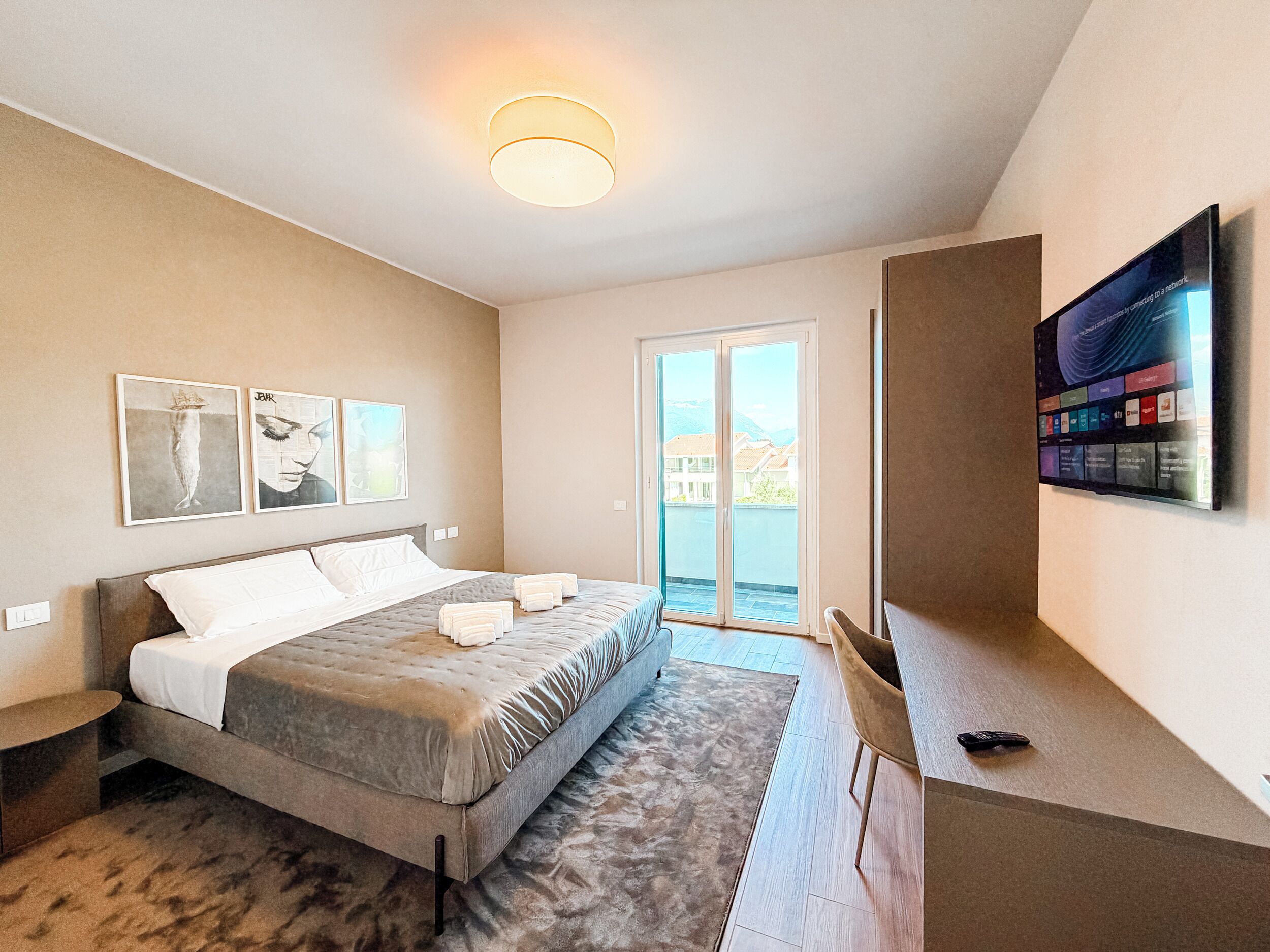 A modern hotel room with a bed, a flat-screen TV, artwork on the wall, and a view through the door.
