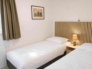 Ein Zimmer mit zwei Einzelbetten, einem gerahmten Bild an der Wand, einer Nachttischlampe und einem Fenster mit Vorhängen.
